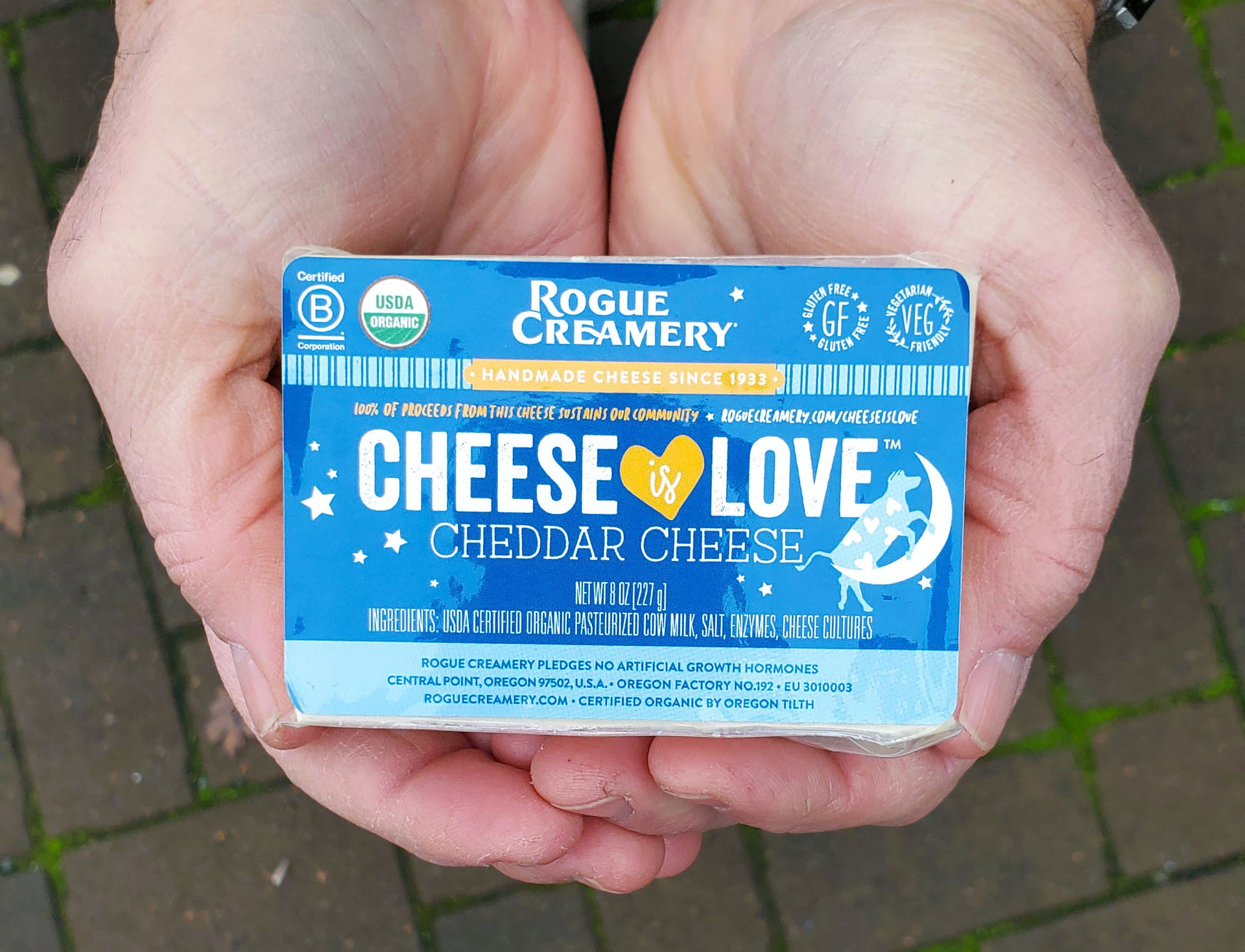 Hands holding a block of Rogue Creamery "Cheese is Love" cheddar cheese with blue packaging and certification labels.