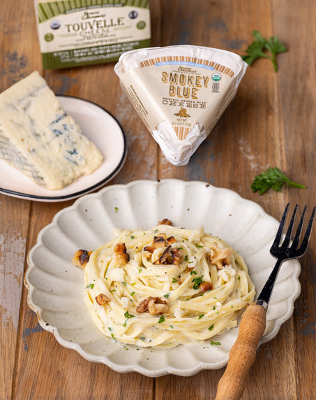 A plate of creamy pasta garnished with walnuts and herbs, accompanied by wedges of Rogue Creamery’s Smokey Blue and Touvelle cheeses on a wooden table.