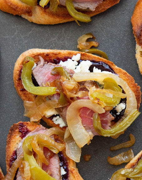 Toasted crostini topped with sliced meat, caramelized onions, green peppers, and crumbled cheese on a dark surface.