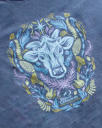 A heather blue t-shirt featuring a cow’s head surrounded by colorful Southern Oregon flora, with “Rogue Creamery” printed below.