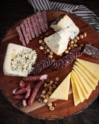 A beautifully arranged wooden board featuring a gourmet selection of sliced and crumbled cheeses, cured meats, and roasted hazelnuts, styled on a striped cloth for a rustic and elegant presentation.