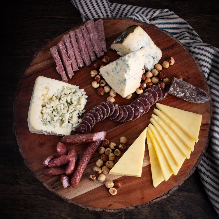 A beautifully arranged wooden board featuring a gourmet selection of sliced and crumbled cheeses, cured meats, and roasted hazelnuts, styled on a striped cloth for a rustic and elegant presentation.