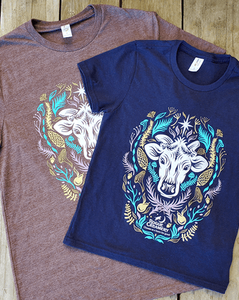 Two t-shirts, one navy and one heather grey-brown, each featuring a cow’s head surrounded by colorful Southern Oregon flora, with “Rogue Creamery” printed below.