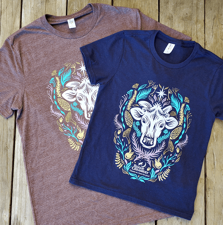 Two t-shirts, one navy and one heather grey-brown, each featuring a cow’s head surrounded by colorful Southern Oregon flora, with “Rogue Creamery” printed below.