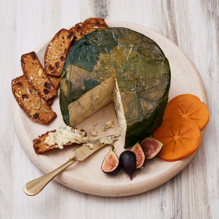 A round cheese wrapped in green leaves on a wooden board, served with fig slices, persimmon slices, and nut-studded crackers with cheese spread.