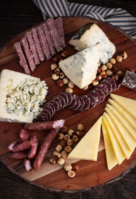 A beautifully arranged wooden board featuring a gourmet selection of sliced and crumbled cheeses, cured meats, and roasted hazelnuts, styled on a striped cloth for a rustic and elegant presentation.