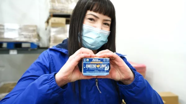 Rogue Creamery Employee makes a heart shape with hands while holding a block of Cheese is Love 8oz extra aged cheddar